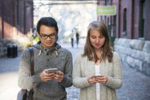 Two young people looking into smartphones while walking on city street