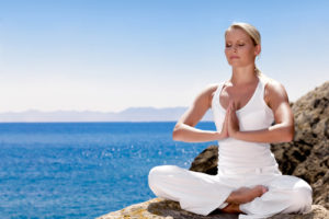 Beautiful positive girl clothing in white sit at the seaside on the rock and meditating in yoga pose