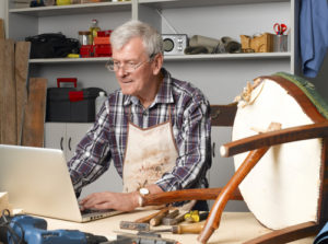 Portrait of retired manual worker sittiing in his small workshop in front of laptop and making online order. Small business.